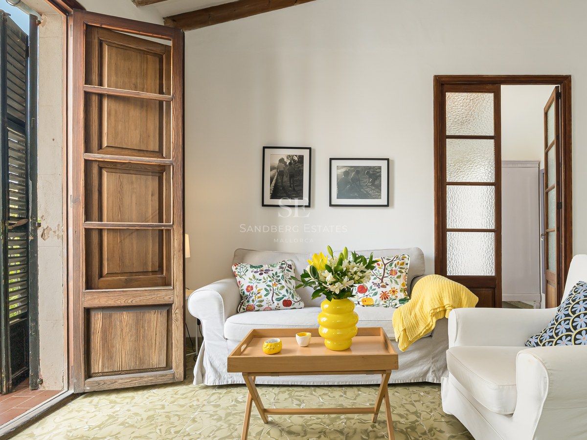 Salón luminoso con sofás blancos, cojines coloridos y grandes puertas de madera que dan a un balcón.