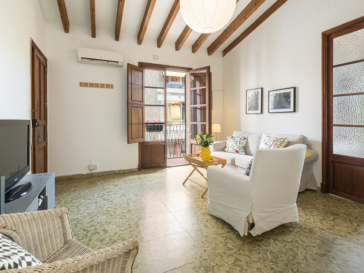 Salón con techos de vigas de madera, suelo de baldosa verde decorada, sofá blanco y puertas de balcón abiertas.