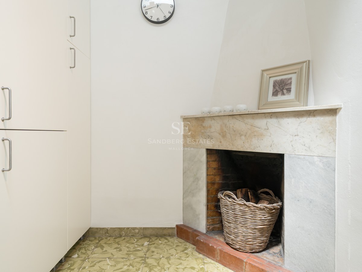 Rincón interior con chimenea de mármol, cesta de mimbre con leña y armario blanco alto.