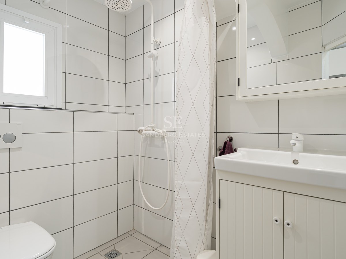 Baño alicatado en blanco con lavabo, inodoro y zona de ducha con cortina blanca.
