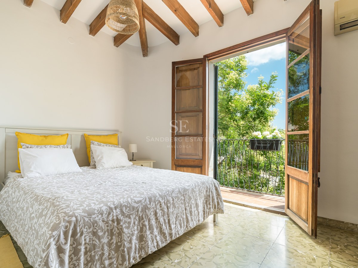 Un dormitorio luminoso con vigas de madera vista y puertas francesas que dan a un balcón soleado con árboles.