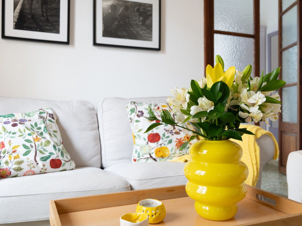 Sala de estar luminosa con sofá blanco, cojines florales y un jarrón amarillo vibrante sobre mesa de madera.