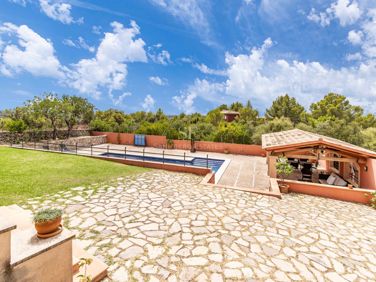 Grand patio en pierre avec piscine, pelouse verte et salon couvert avec poutres en bois et toit en tuiles.