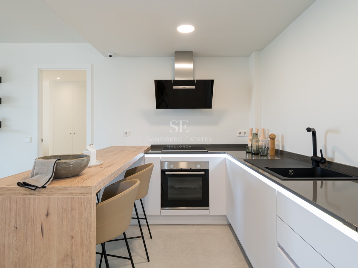 Minimalist white kitchen featuring a light oak breakfast bar, black sink, and sleek integrated appliances.