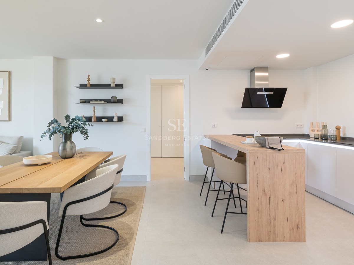 Minimalist white kitchen with wood island and adjacent dining area featuring designer chairs and light stone flooring.
