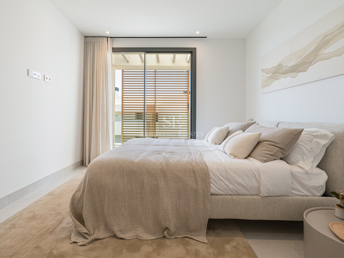 Bright modern bedroom with a beige upholstered bed, abstract wall art, and glass doors opening to a sunny terrace.