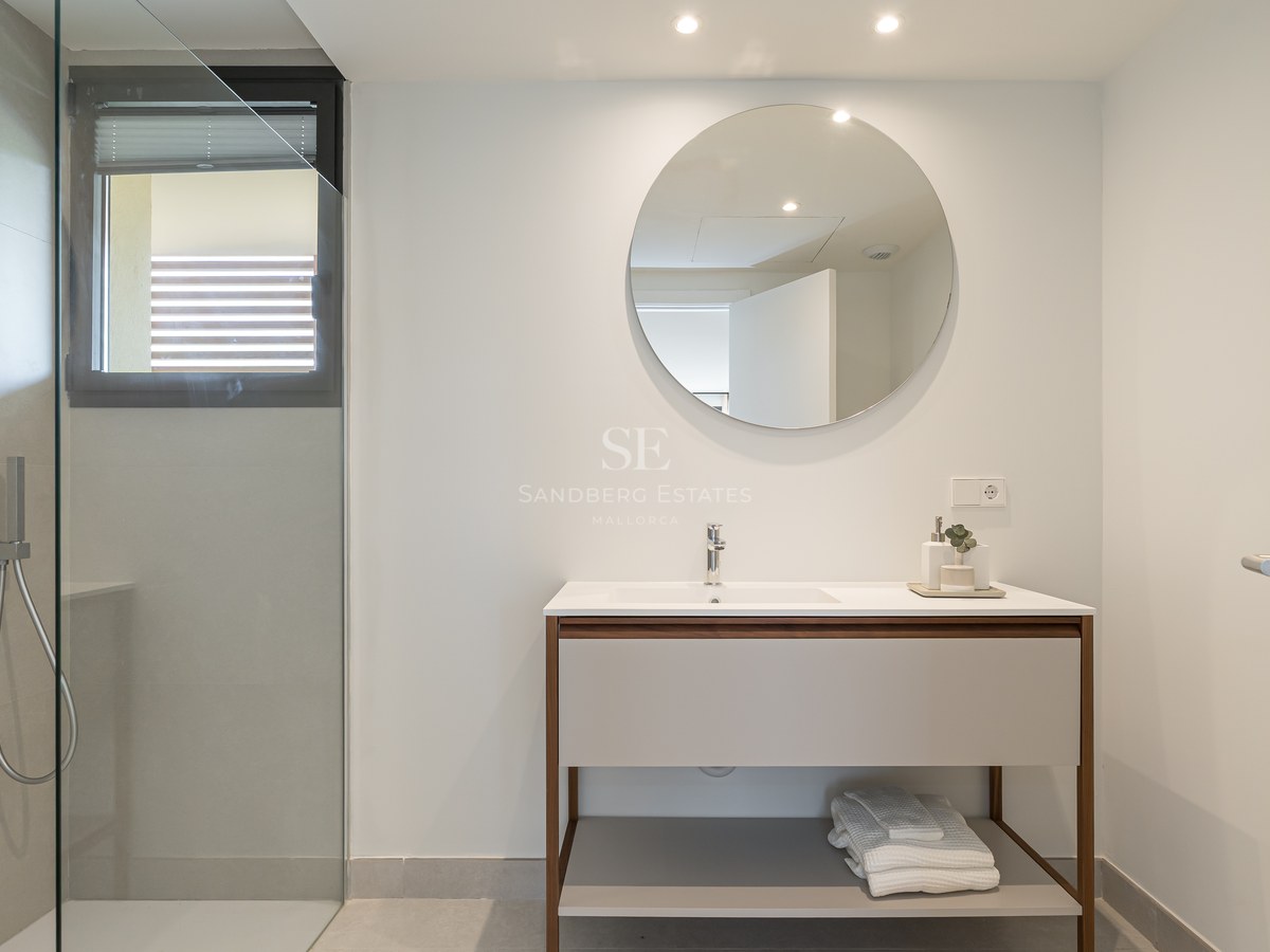 A modern bathroom featuring a walk-in glass shower, a circular mirror over a wooden vanity, and grey tiled walls.