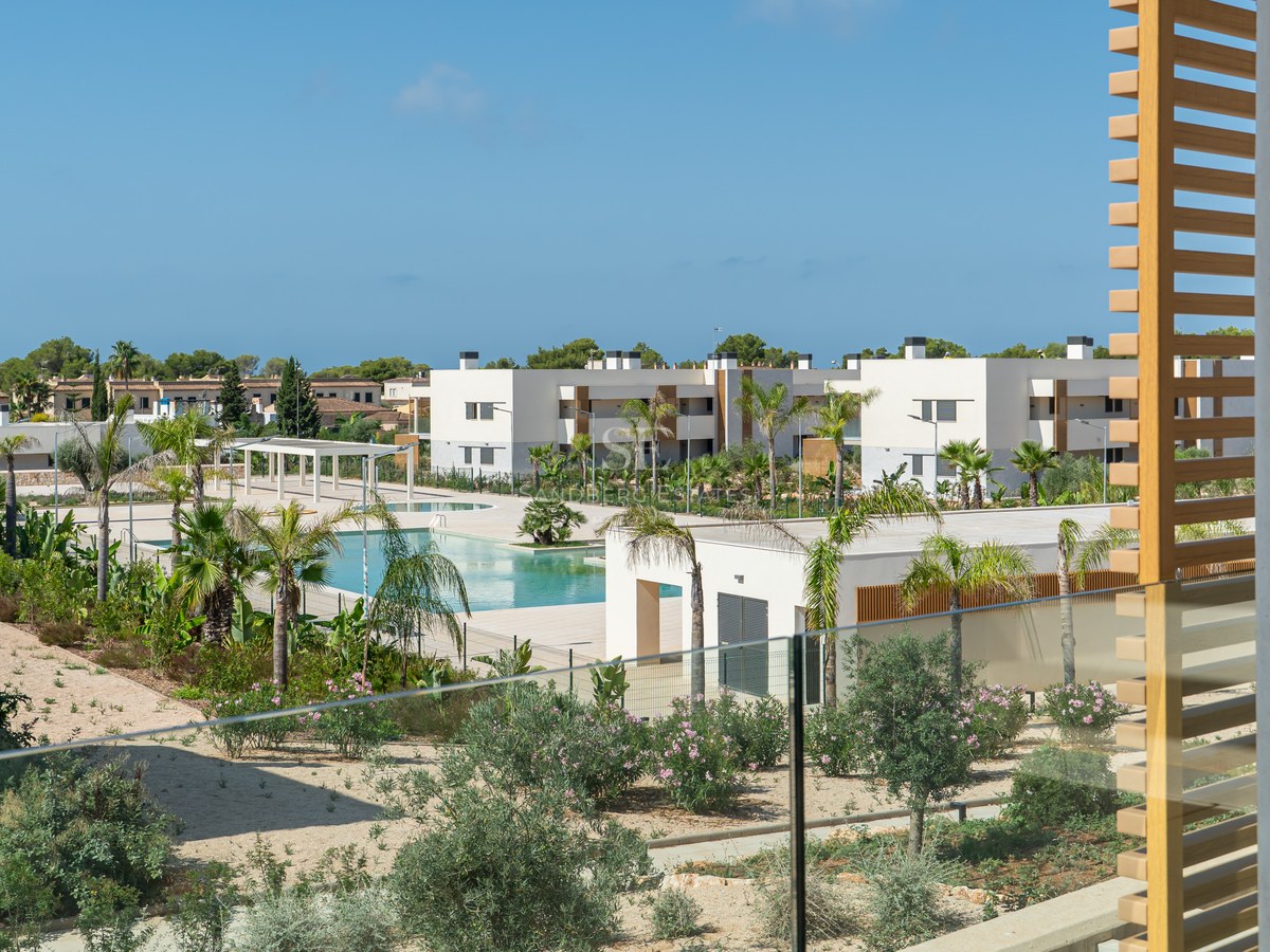 View from a modern terrace with glass railing overlooking a large turquoise swimming pool, palm trees, and white buildings.