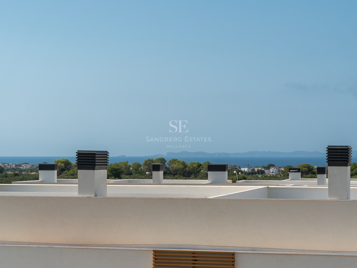 View from a modern white rooftop looking out over pine trees towards the blue Mediterranean Sea and a distant island.