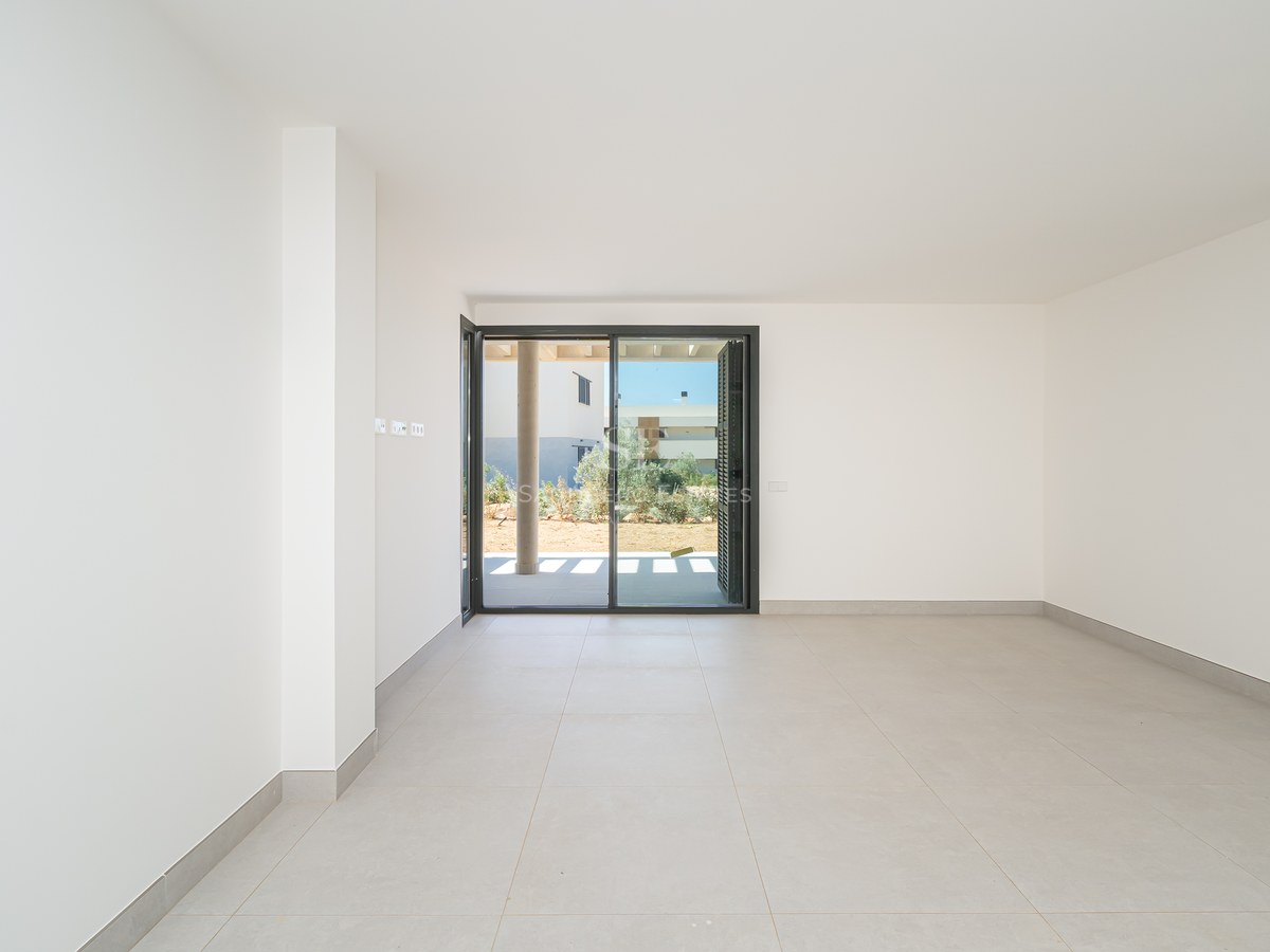 Empty modern room with white walls, large grey floor tiles, and a sliding glass door opening to a garden.