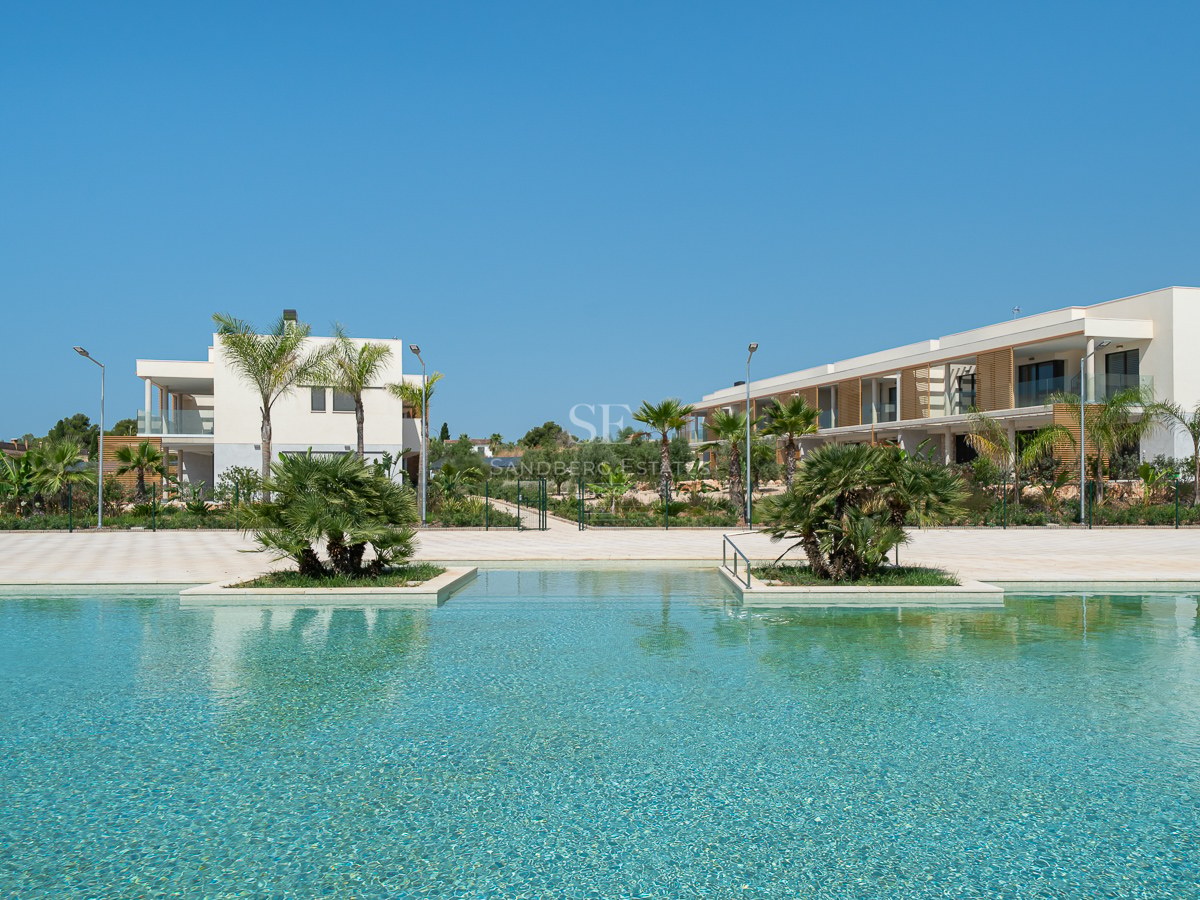 Large turquoise swimming pool in front of modern white villas under a clear blue sky with palm trees.