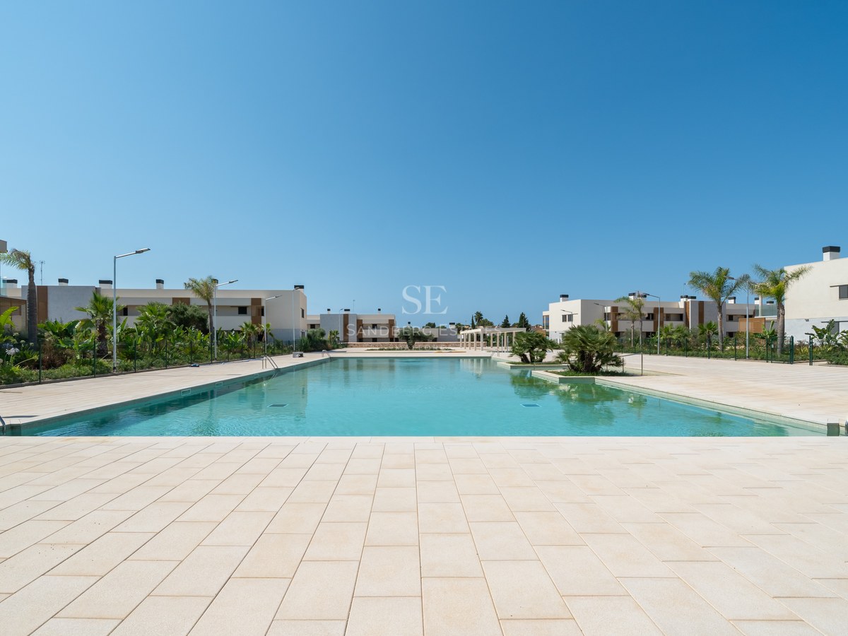 Large turquoise swimming pool surrounded by light stone paving and modern white villas under a clear blue sky.