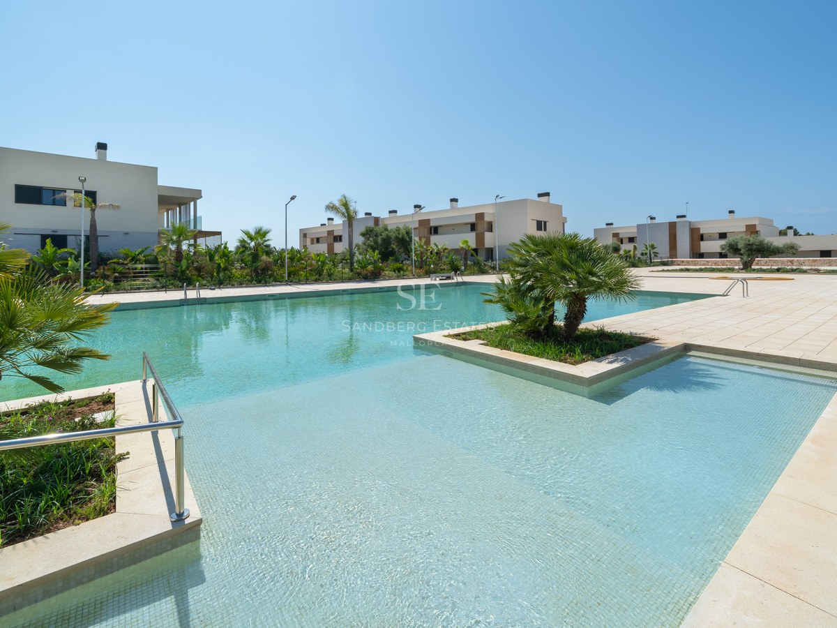 Spacious turquoise swimming pool surrounded by stone tiling and contemporary white buildings under a clear blue sky.