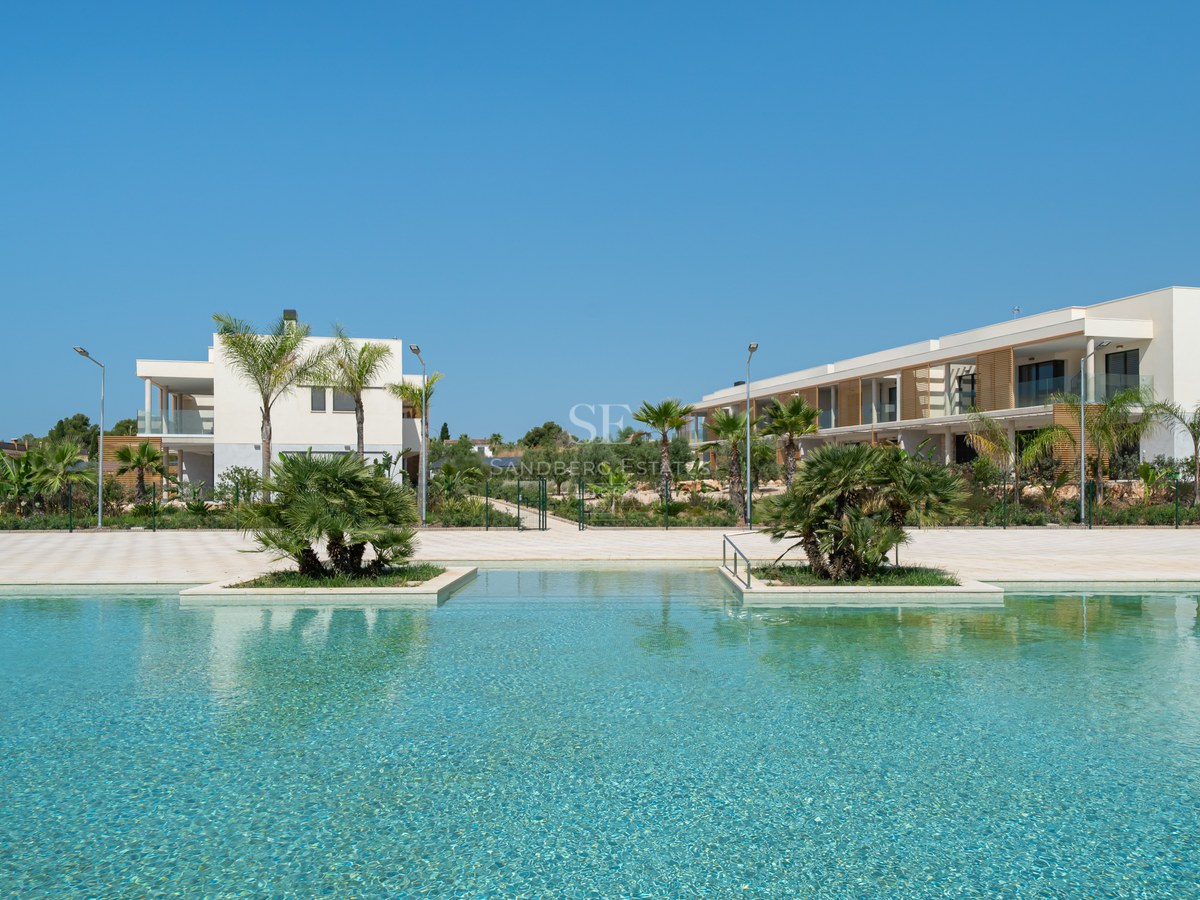 Large turquoise swimming pool with palm tree islands in front of white modern Mediterranean buildings under a blue sky.