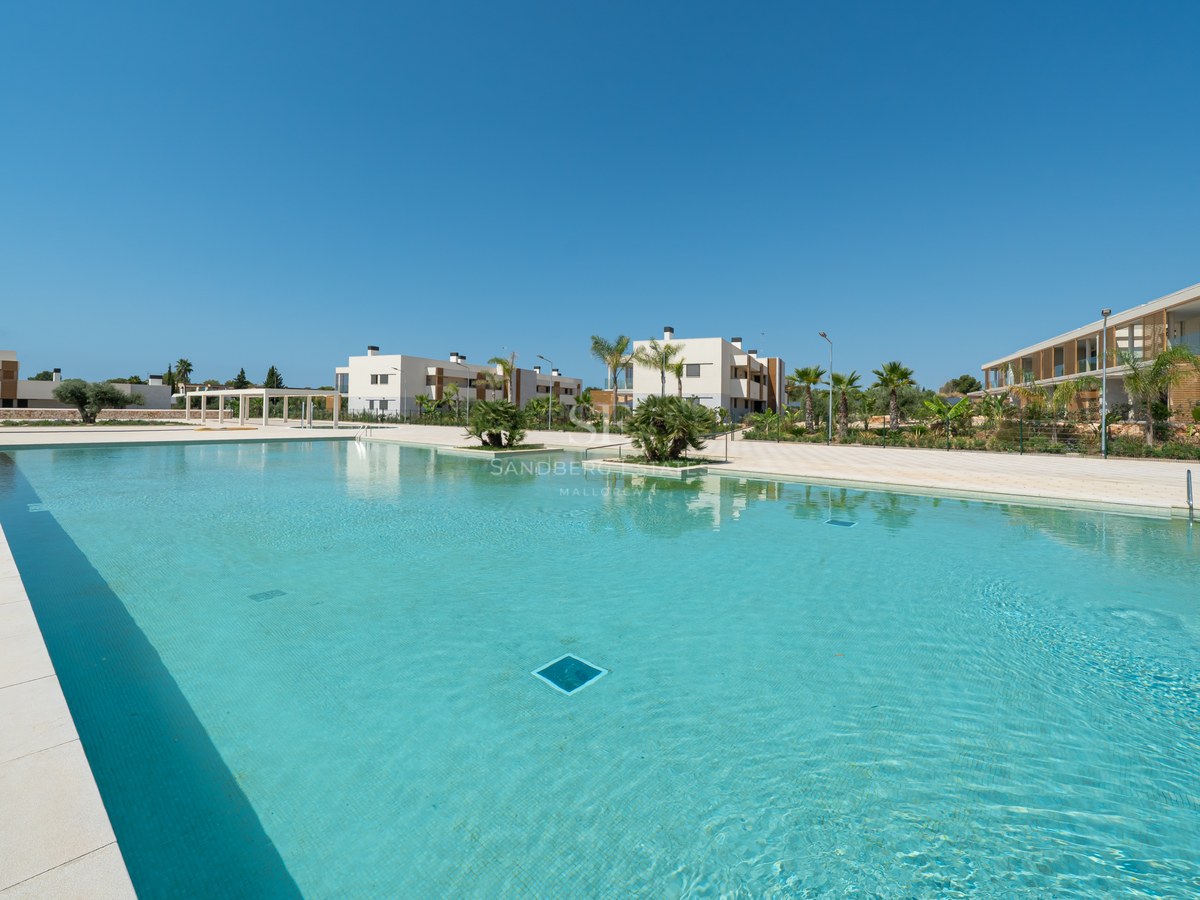 Large turquoise outdoor pool surrounded by cream stone tiles and modern white apartment buildings under a clear blue sky.