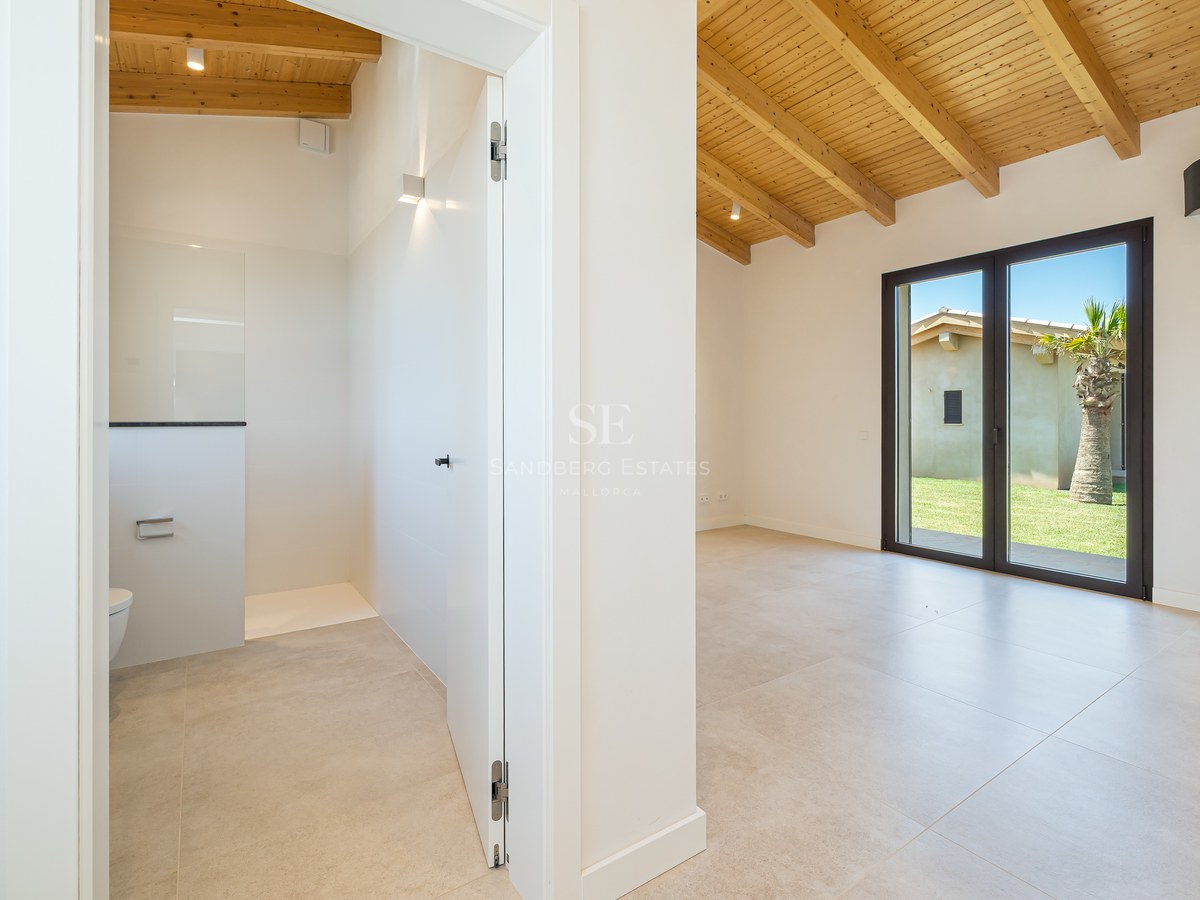 Chambre lumineuse avec poutres en bois clair, carrelage grand format et aperçu de la salle de bain attenante.