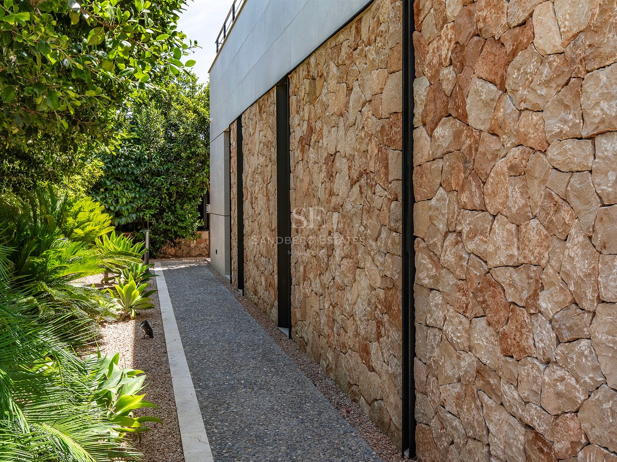 A textured natural stone wall adjacent to a gravel path and lush Mediterranean greenery under bright daylight.