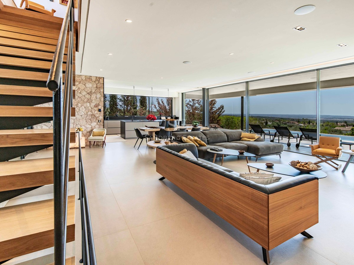 Spacious modern living room with wooden stairs, stone wall, and floor-to-ceiling windows overlooking a terrace and landscape.