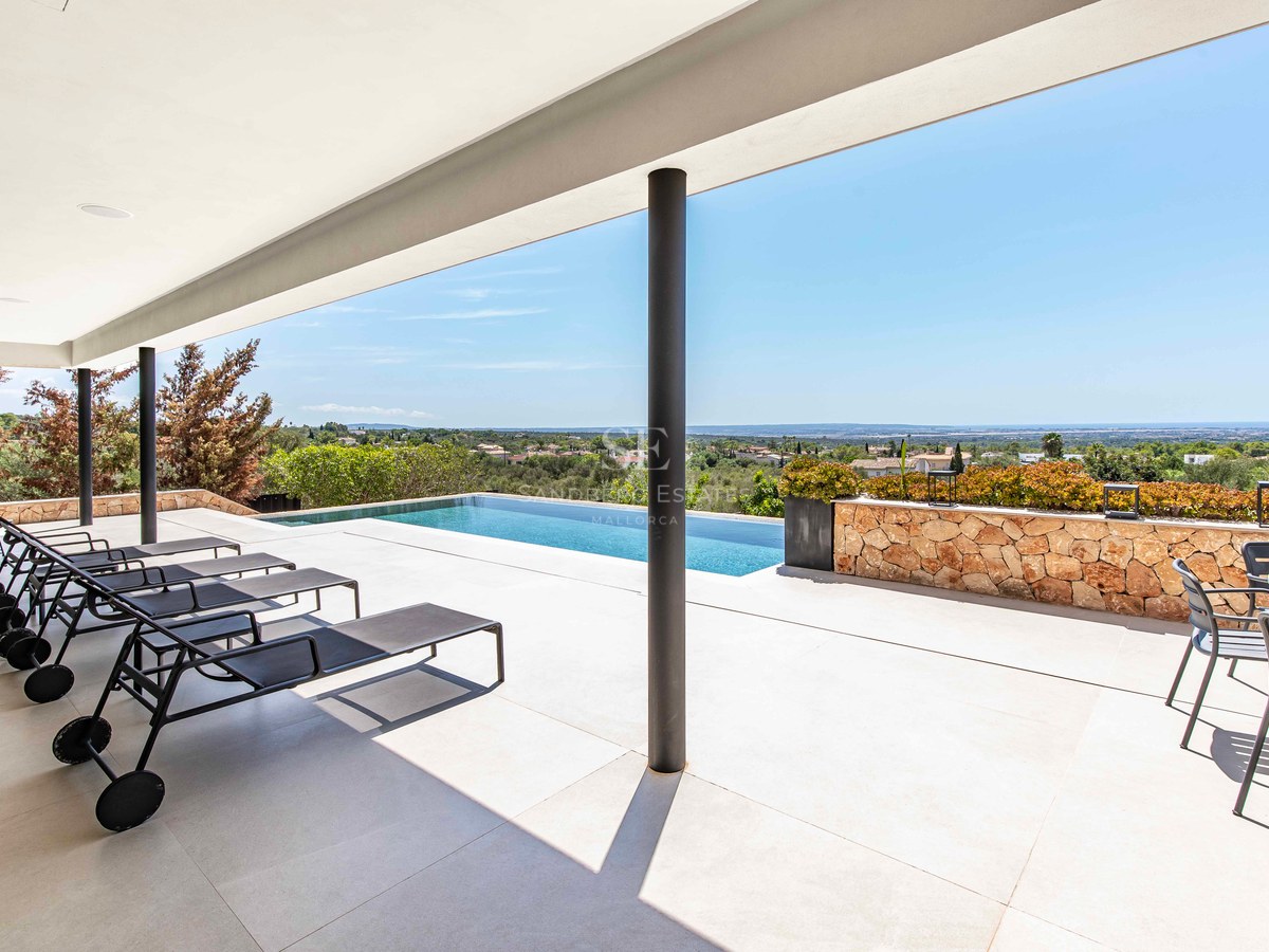 A spacious modern white tiled terrace with black sun loungers, an infinity pool, and a view of the landscape and sea.