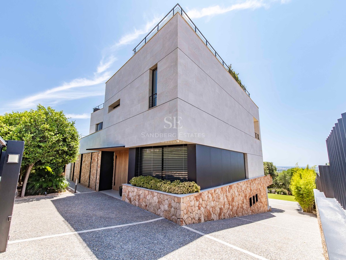 Modern villa exterior featuring natural stone walls, wooden entrance, and a gravel driveway under a clear blue sky.