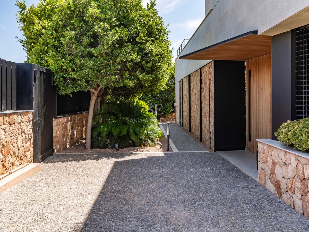 A contemporary villa entrance featuring a wooden door, stone walls, a gravel path, and lush green trees under bright sun.