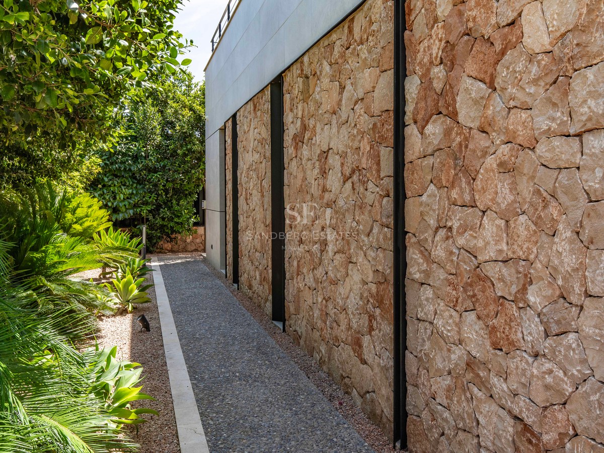 Side view of a contemporary villa's stone facade with a gravel path and lush green Mediterranean plants.