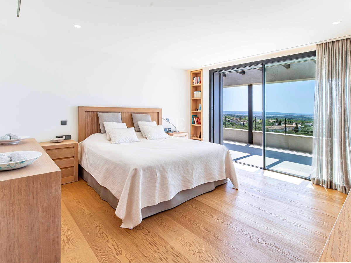 Bright modern bedroom with light oak floors and floor-to-ceiling glass doors opening to a sunny terrace and sea views.