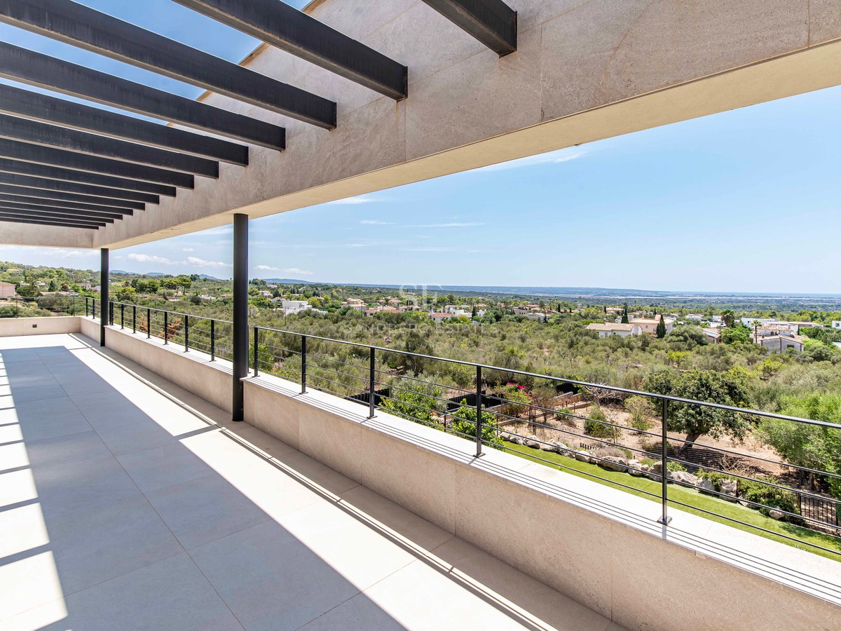 Wide modern terrace with a black slat pergola overlooking a green valley and distant coastline under a clear blue sky.