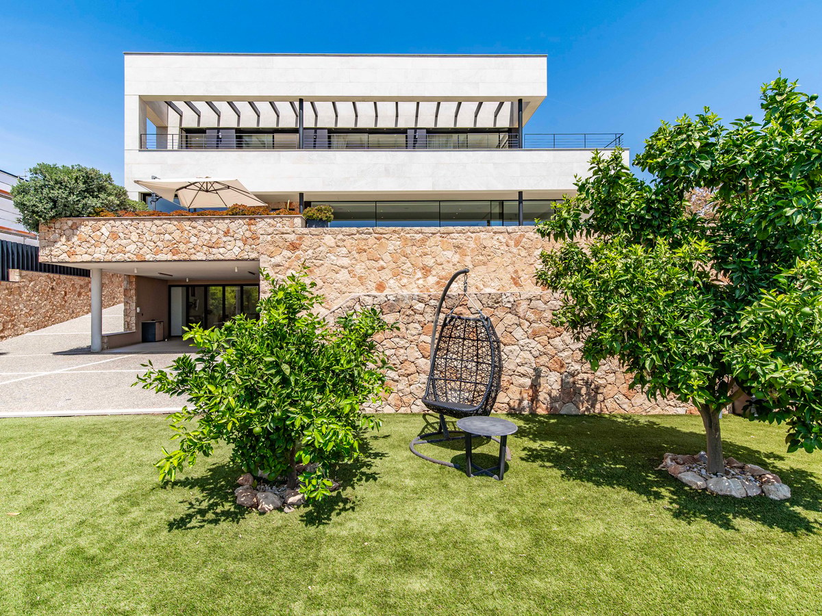 Exterior view of a modern three-level villa featuring stone walls, white upper levels, glass balconies, and a green lawn.