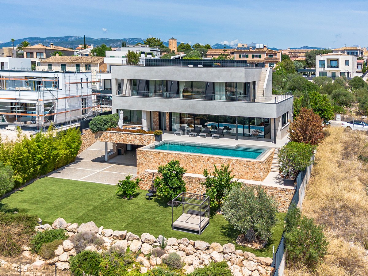 Aerial view of a contemporary three-story villa with a turquoise pool, natural stone walls, and a manicured lawn garden.