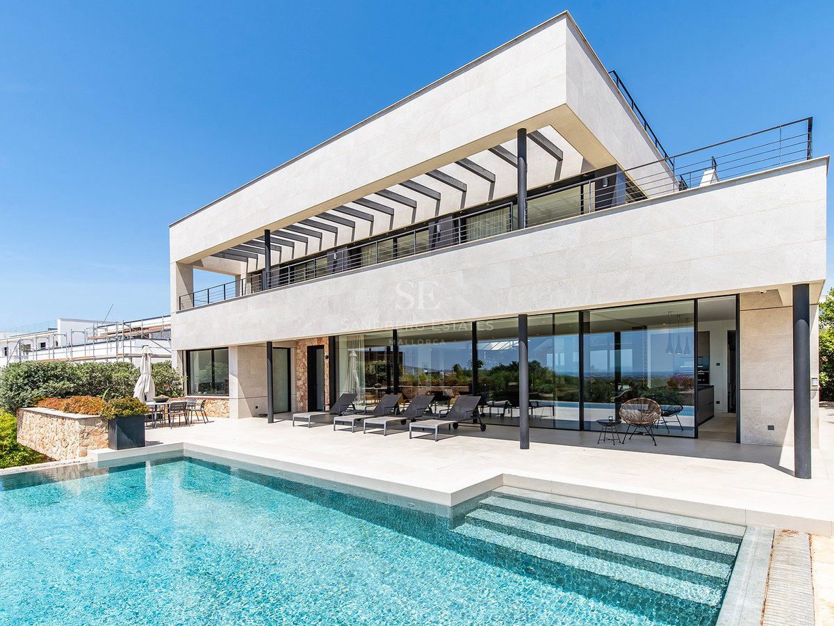 Contemporary two-story villa with natural stone facade, large swimming pool, and sun loungers under a clear blue sky.