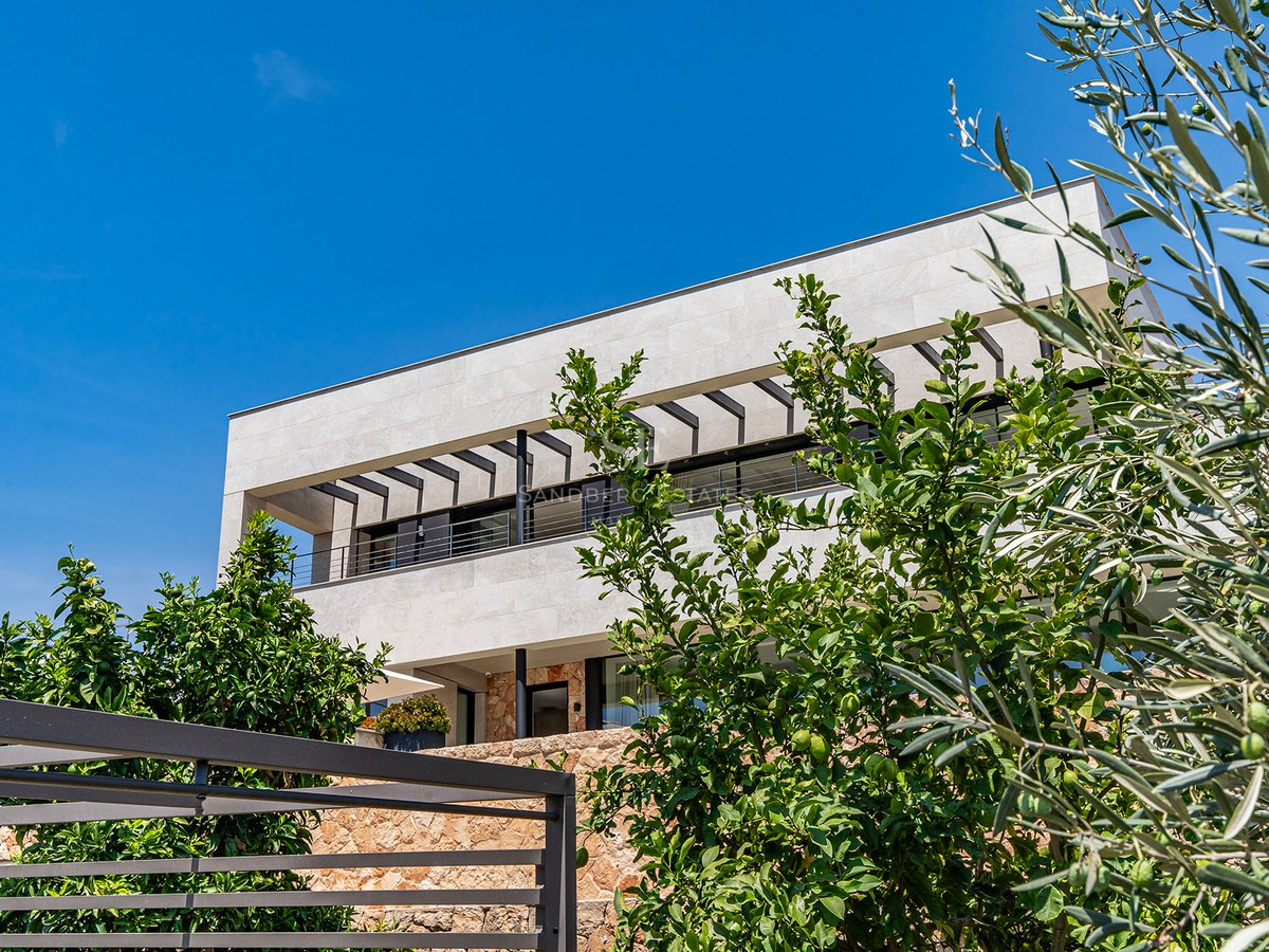 Modern two-story villa with stone facade, glass balconies, and lush olive and citrus trees under a clear blue sky.