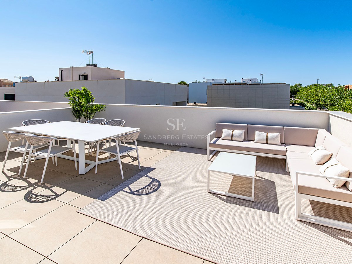 Terraza soleada con mesa de comedor blanca, sillas de cuerda y sofá rinconero beige sobre alfombra de exterior.