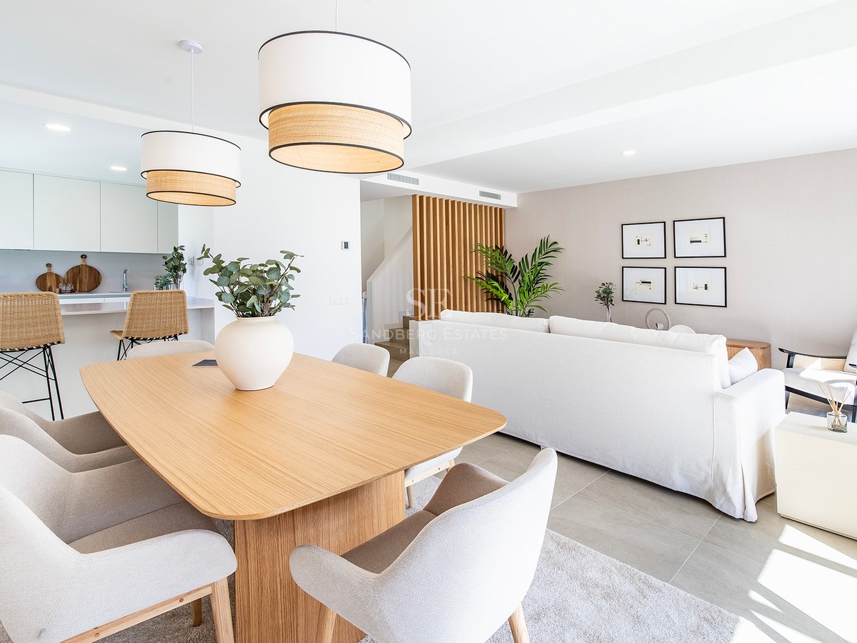 Salón comedor luminoso con mesa de madera clara, sillas beige y sofá blanco bajo luz natural.