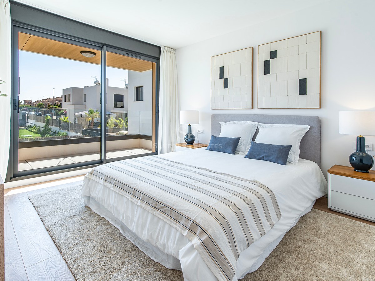 Dormitorio moderno luminoso con cama doble, suelos de madera y ventanales de suelo a techo hacia una terraza.