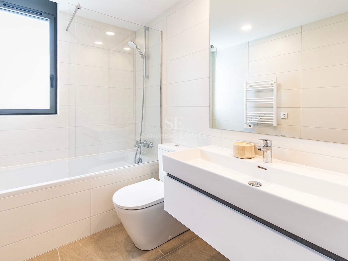 Cuarto de baño blanco con bañera, mampara de cristal, lavabo suspendido y suelo de gres beige.