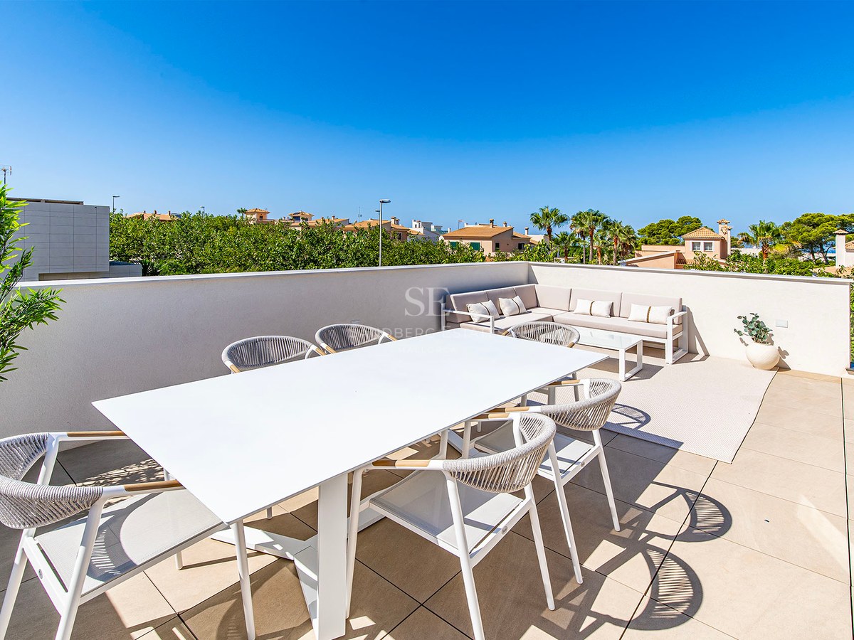 Terraza luminosa en la azotea con mesa de comedor blanca y zona de estar bajo un cielo azul despejado.