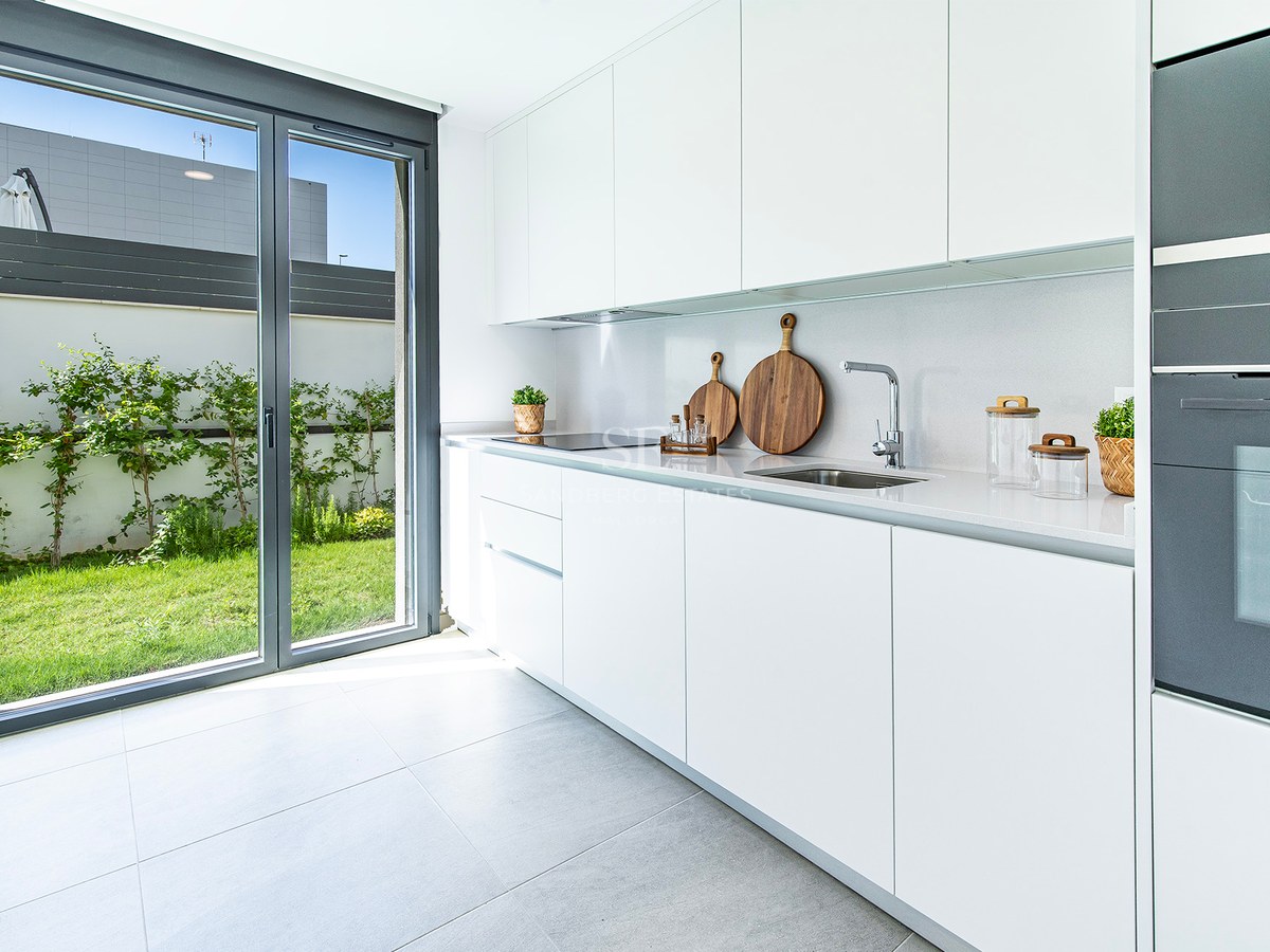 Cocina blanca con armarios sin tiradores, encimera de piedra y puerta acristalada hacia un jardín privado.
