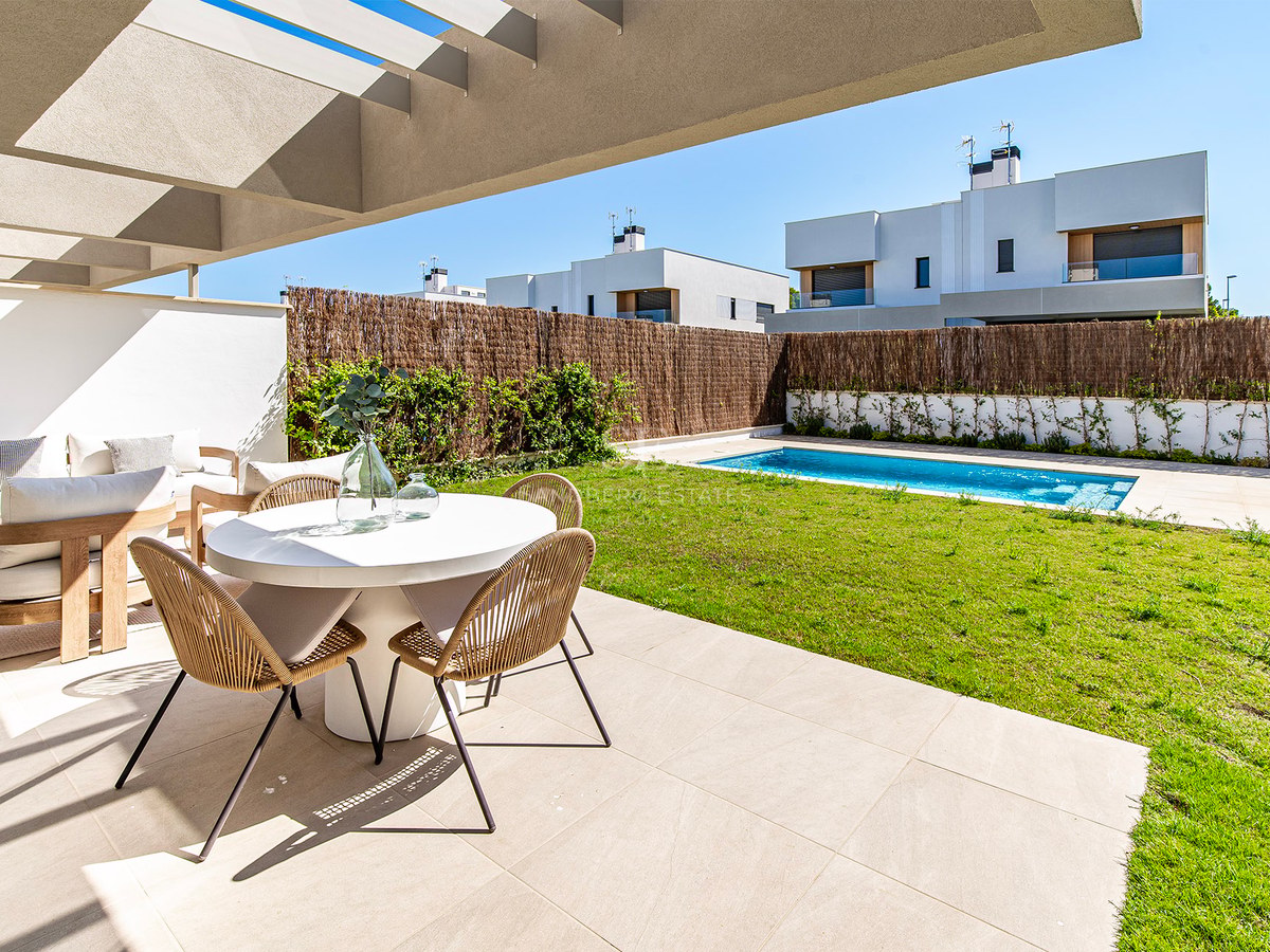 Terraza soleada con mesa de comedor blanca, sillas de mimbre, césped y una piscina rectangular al fondo.
