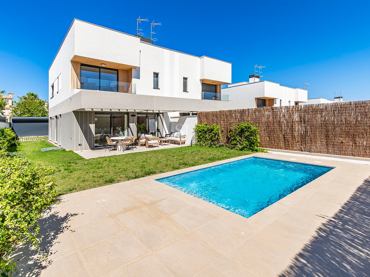 Villa moderna blanca de dos plantas con piscina rectangular, terraza de piedra y césped bajo un cielo azul despejado.