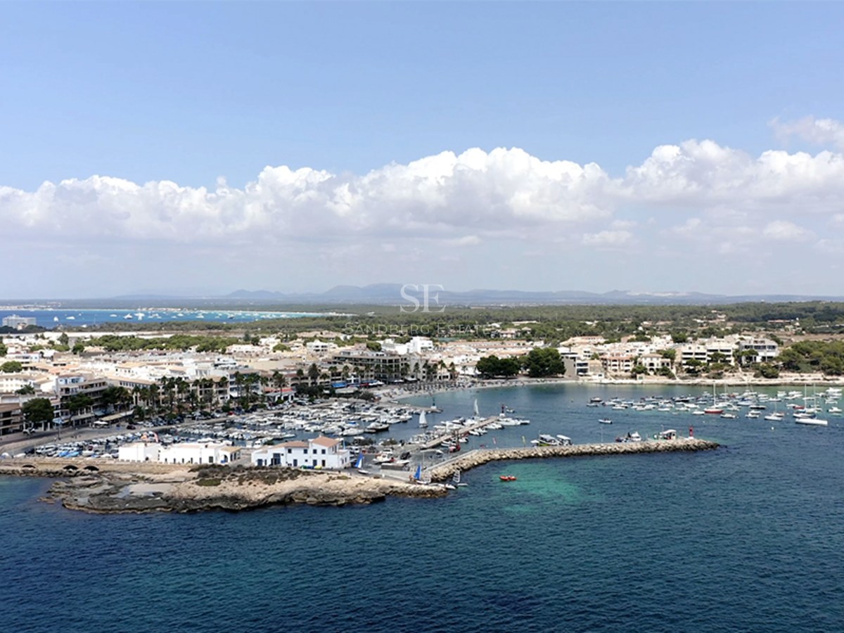 Elevated aerial view of a busy marina with white yachts, coastal buildings, and clear turquoise sea under a blue sky.