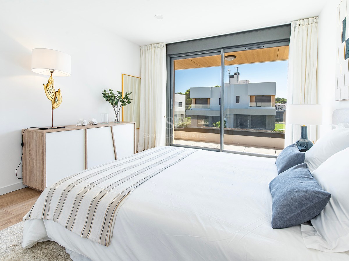 Helles Schlafzimmer mit Doppelbett, Parkettboden und großen Glasschiebetüren zu einer sonnigen Terrasse.