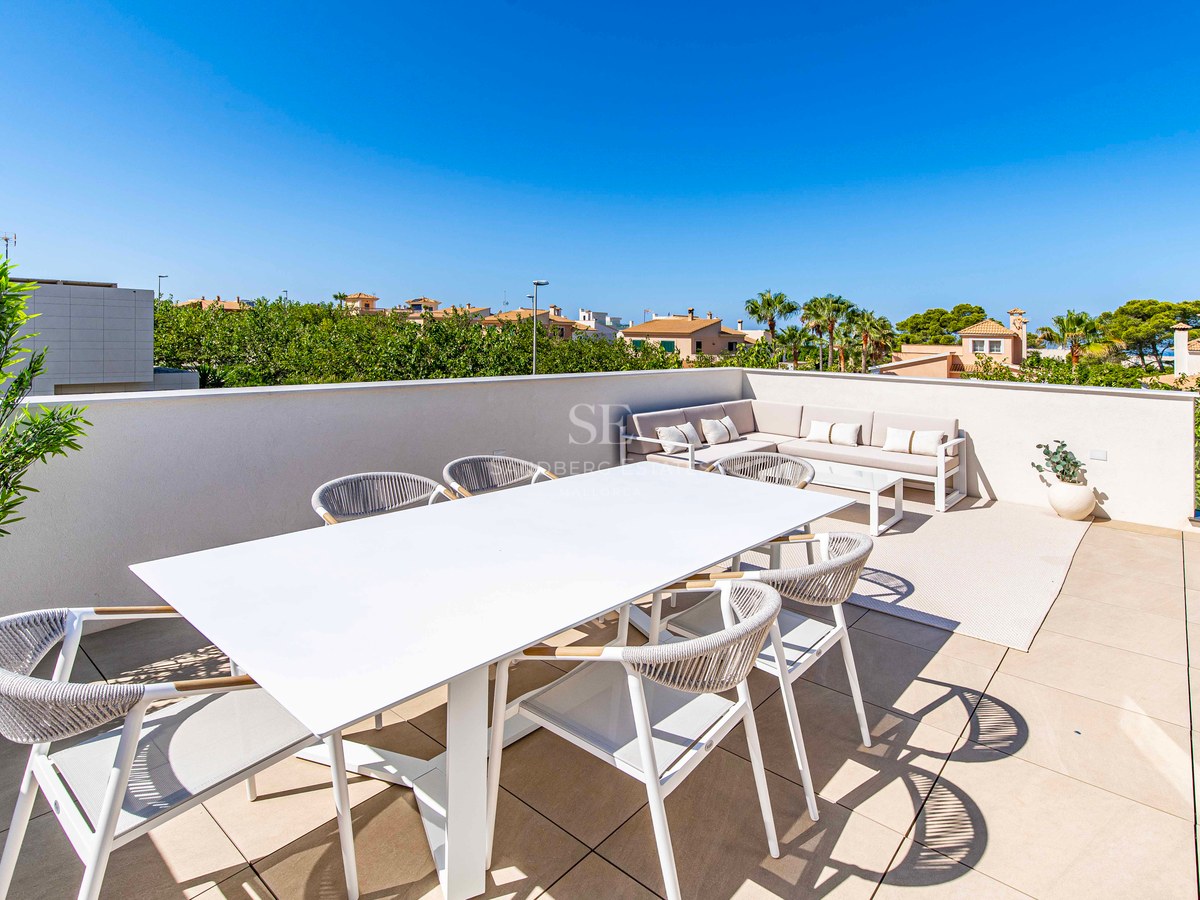 Sonnige Dachterrasse mit weißem Esstisch, Seilstühlen und Ecksofa unter blauem Himmel.