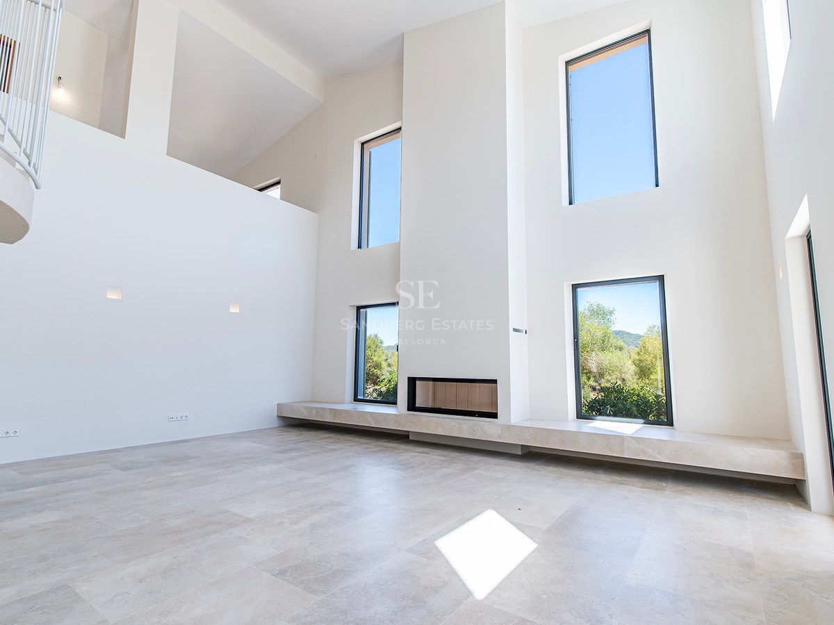 Salón minimalista de doble altura con paredes blancas, grandes ventanales, chimenea integrada y suelos de piedra clara.