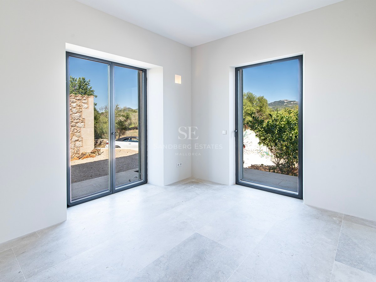 Habitación minimalista vacía con paredes blancas, suelos de piedra gris y dos grandes ventanales con vistas al exterior.