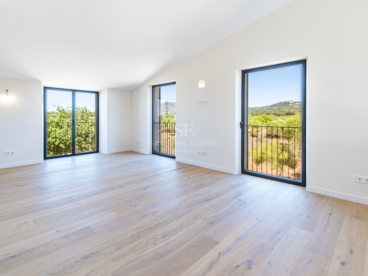Habitación vacía con suelos de madera clara, techos inclinados y grandes ventanales con vistas al paisaje verde.