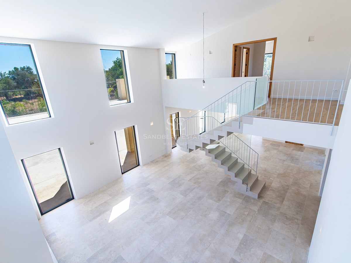 Salón de doble altura con suelos de piedra, escalera moderna y grandes ventanales con luz natural en villa de lujo.