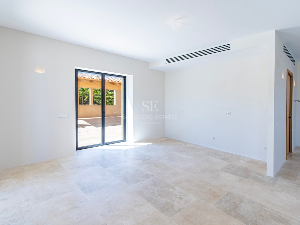 Habitación vacía con suelo de piedra natural crema, paredes blancas y puerta de cristal hacia un patio soleado.