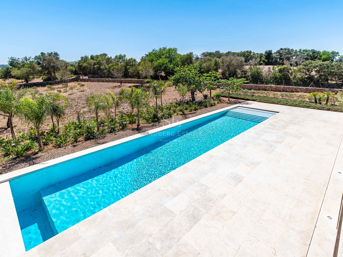 Piscina rectangular turquesa rodeada por una amplia terraza de piedra crema y vegetación mediterránea.
