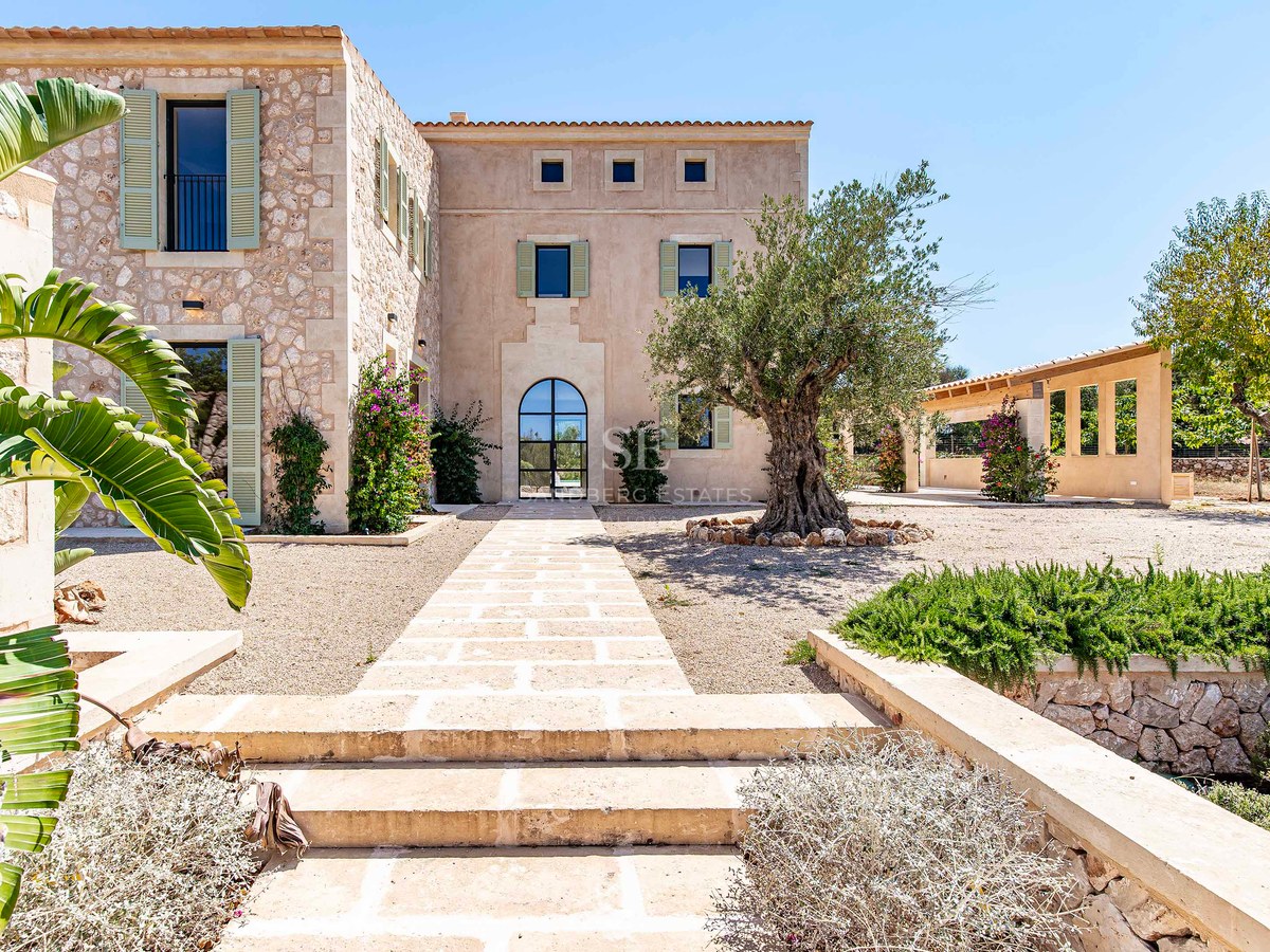 Fachada principal de una villa de lujo en piedra natural con puerta de arco, persianas verdes y un olivo centenario.